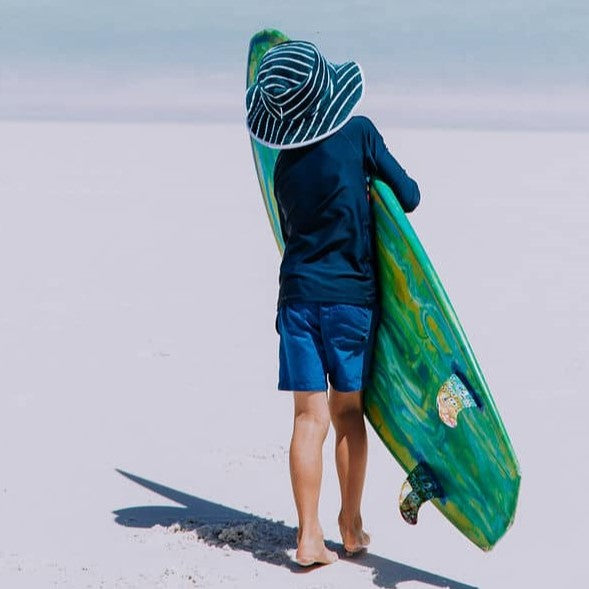 Back view of a boy at the beach in Western Australia carrying a surf board wearing swimwear and Swoodi Beach Bucket Hat