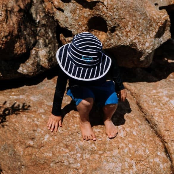 Boy playing on rocks at Wharton Beach wearing swimwear and navy striped Swoodi bucket hat