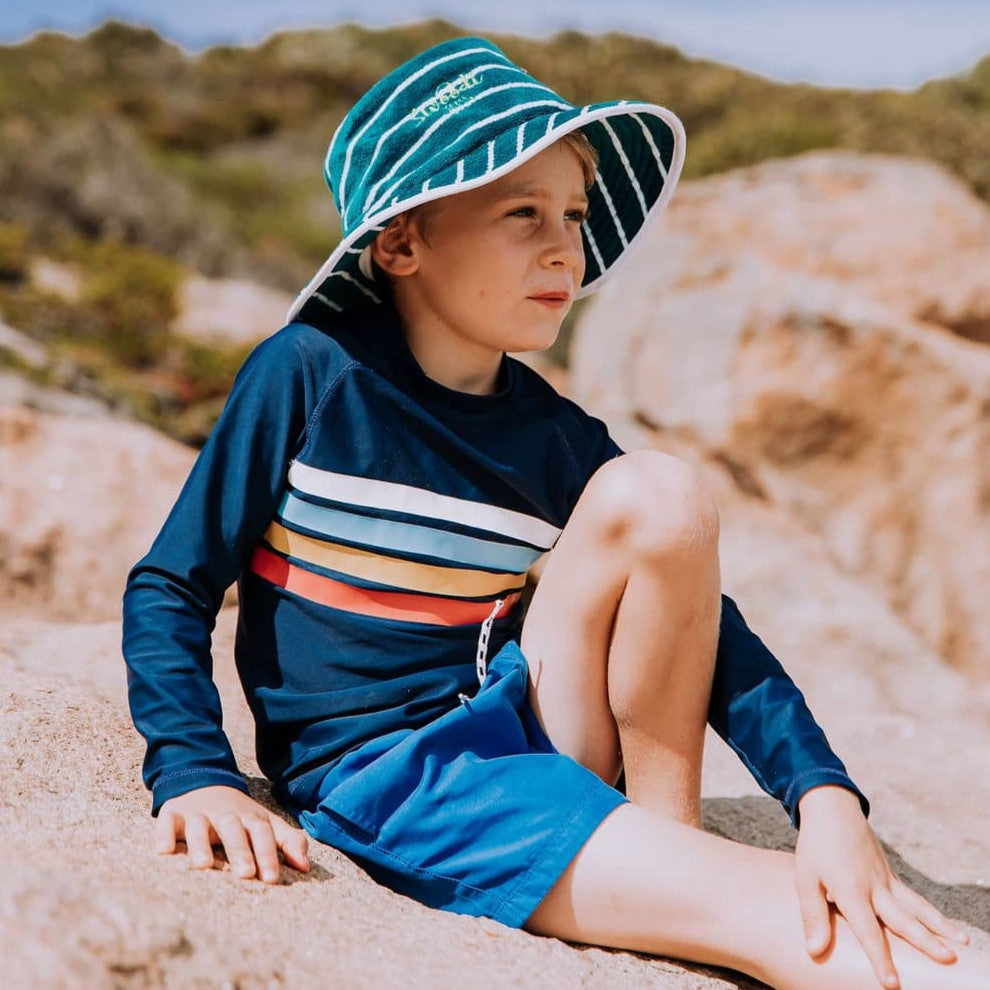 Boy wearing Swoodi wide brimmed toweling bucket hat in turquoise and green, sitting on a rock looking out at the ocean at Wharton Beach