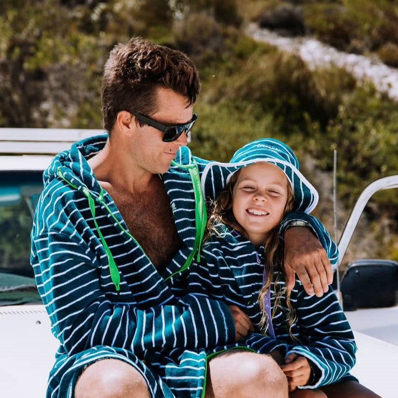 A girl wearing UPF 50+ bucket hat and matching Swoodi zip up hooded towel in turquoise with her Dad, sitting on a land cruiser at the beach