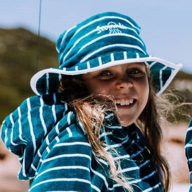 Girl at the beach wearing Swoodi zip up hooded towel with matching bucket hat in turquoise and white stripe