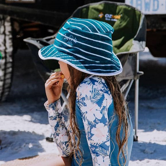 Side view of a girl sitting on the beach wearing Swoodi sun protection bucket hat and swimwear, while eating