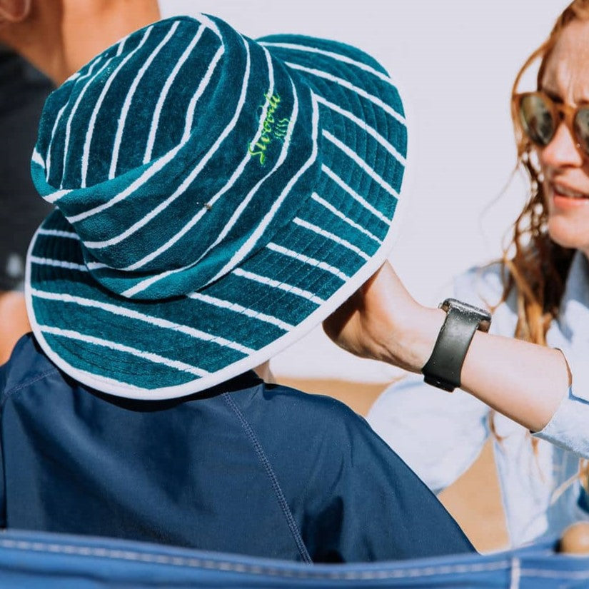 Boy wearing turquoise with green Swoodi Sun Protection Bucket Hat at the beach while his mum puts sunscreen on his face 