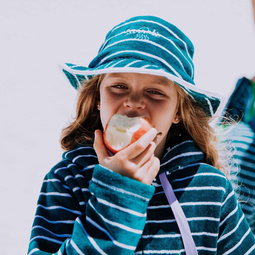 Girl eating an apple at the beach, wearing Swoodi turquoise towel bucket hat with matching zip up swim robe