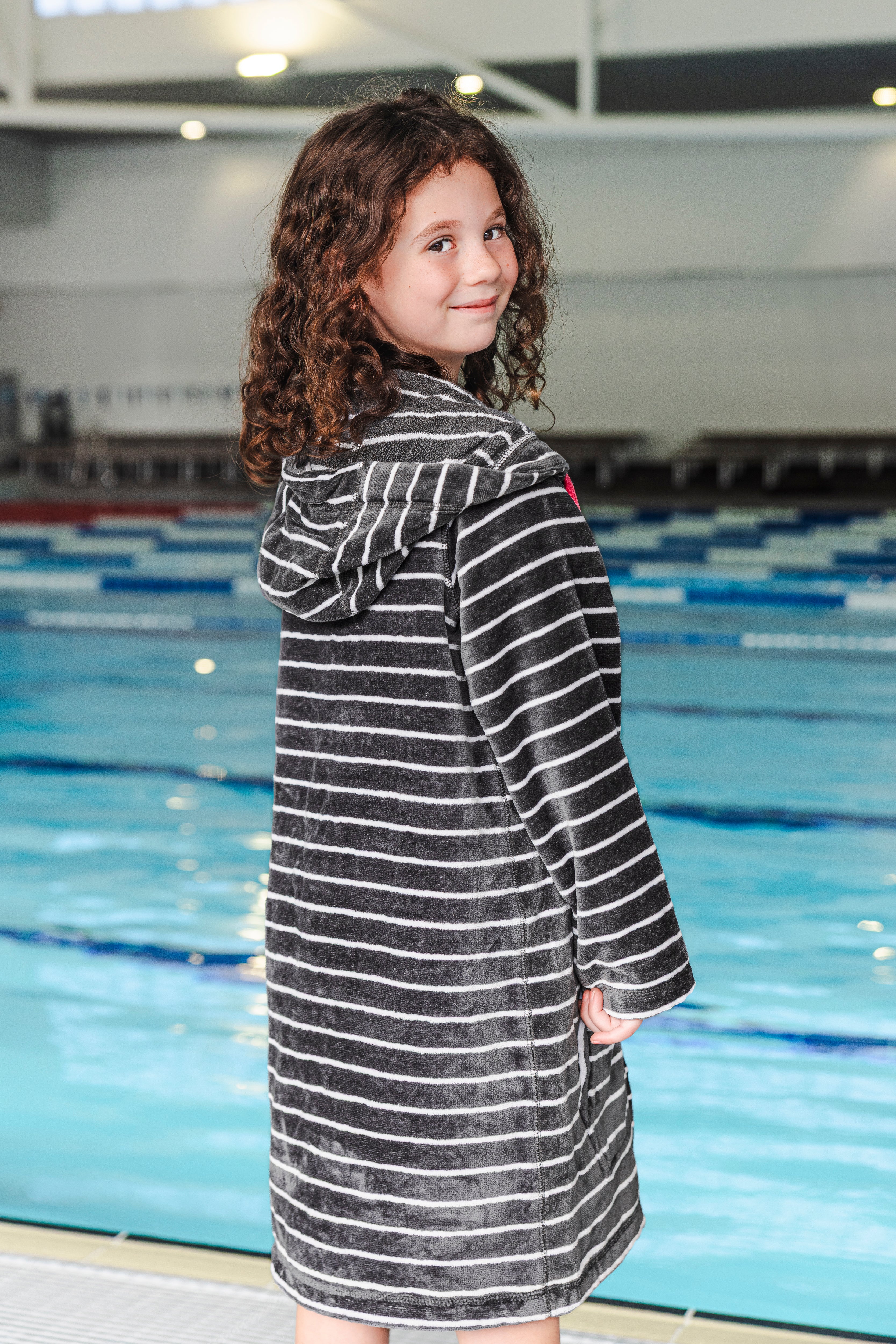 Young girl in a striped zip up hooded towel standing by a pool looking over her shoulder