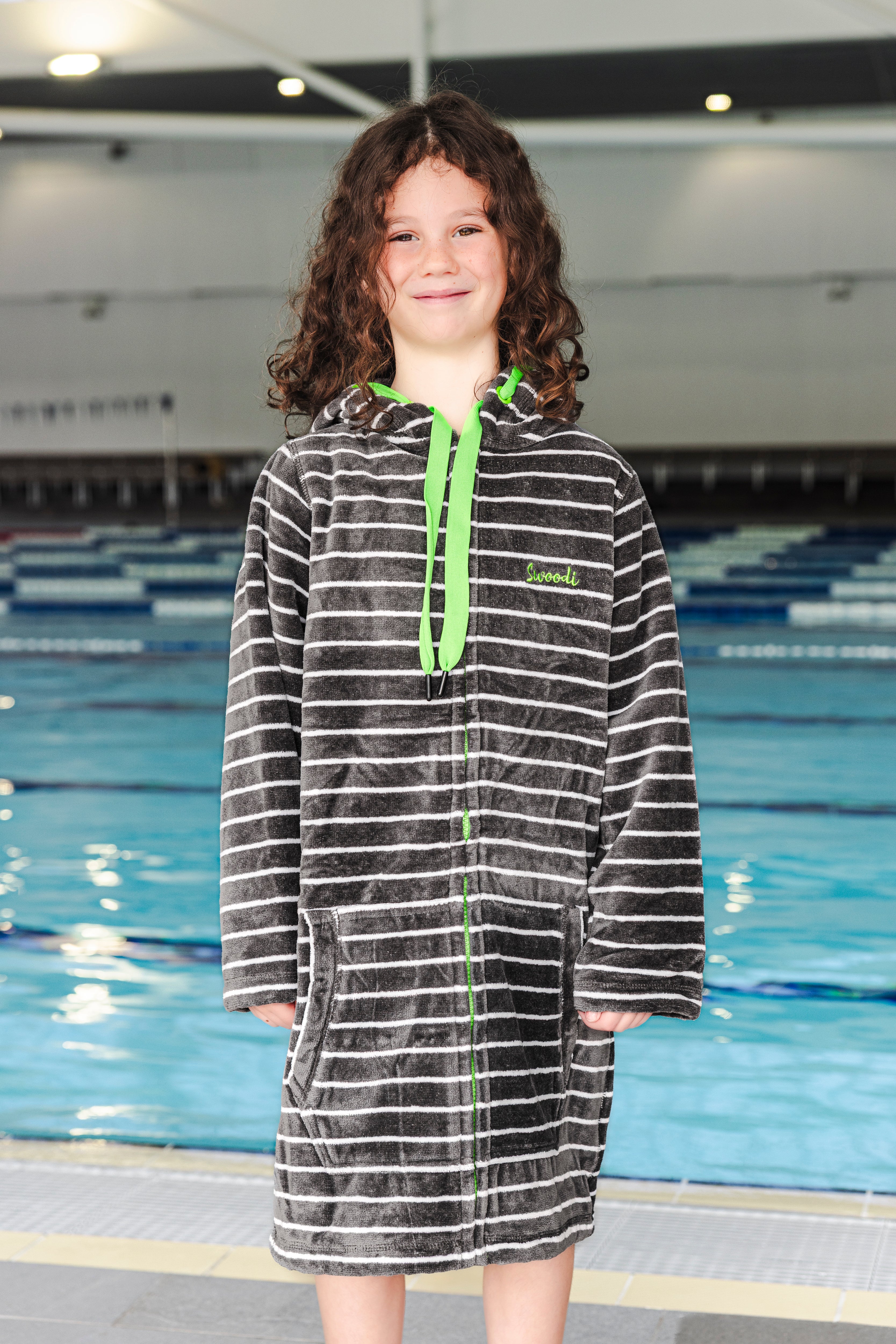 Child wearing a striped hooded towel with a green zipper in an indoor swimming pool setting