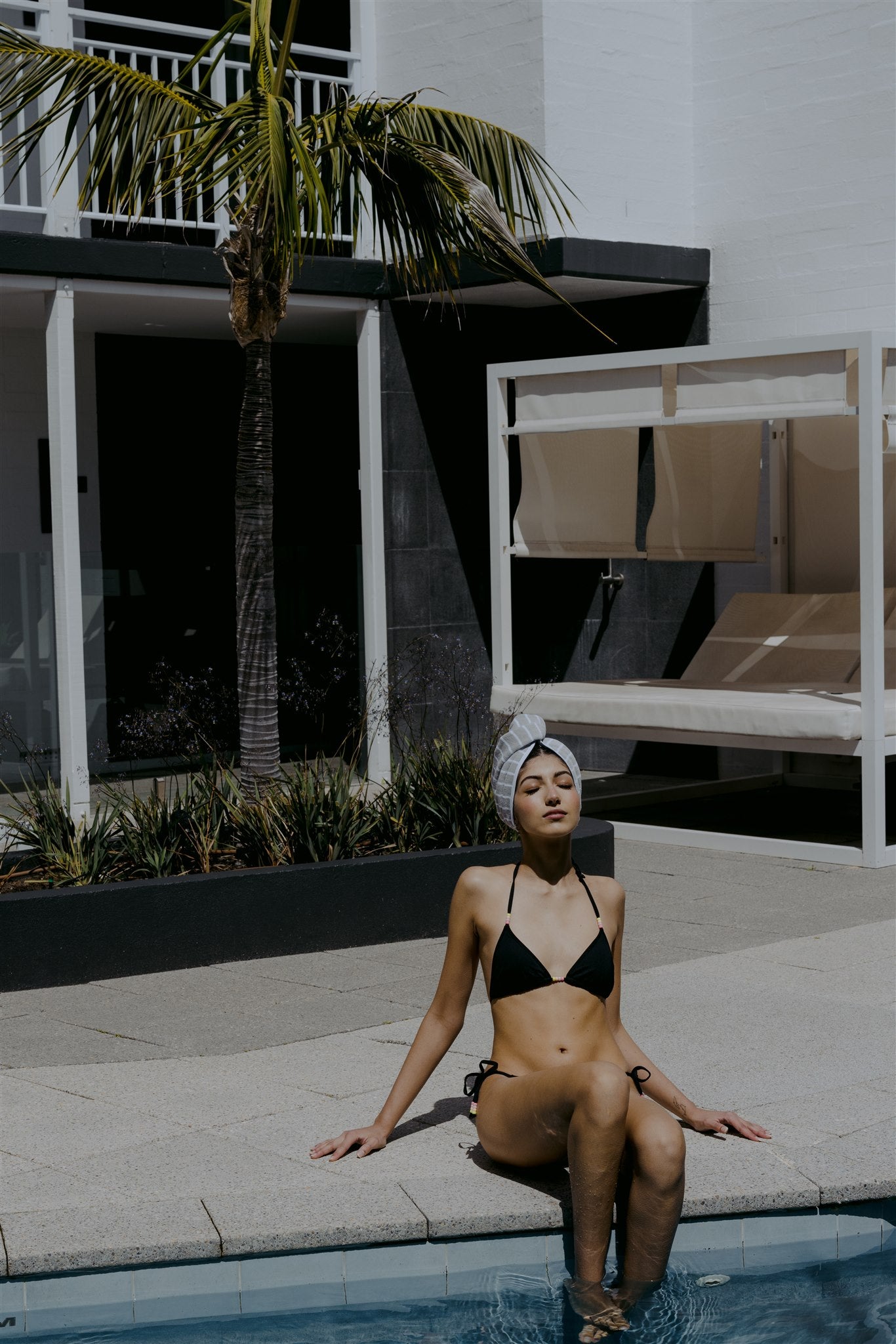 Female model sitting on edge of pool wearing a black bikini and hair wrap with palm tree in the background 